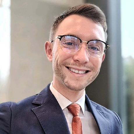 Uomo sorridente con occhiali, giacca blu e cravatta arancione, sfondo sfocato.