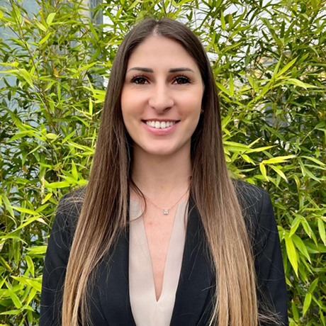 Donna sorridente con capelli lisci lunghi, giacca nera e camicia beige, davanti a un cespuglio verde.